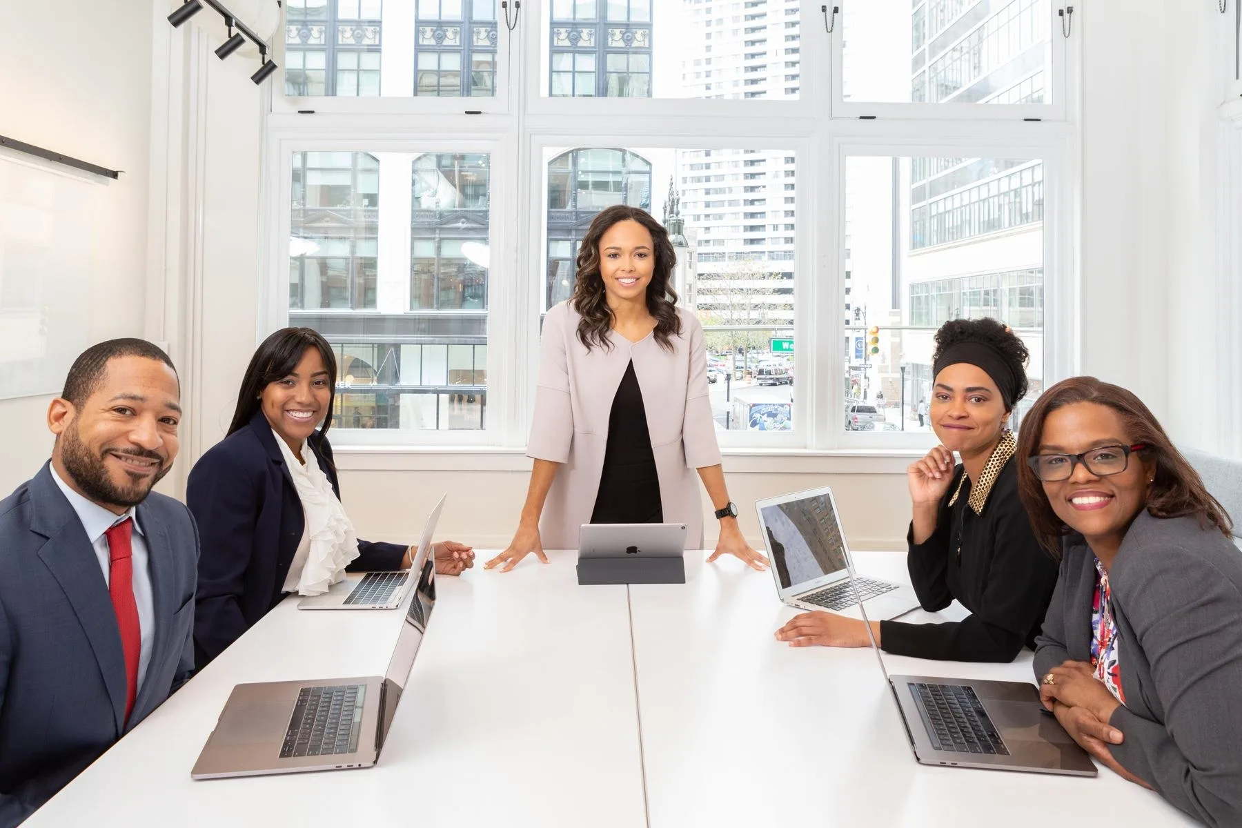 Five colleagues smiling around a bright meeting-room table with laptops, one standing at the head of the table