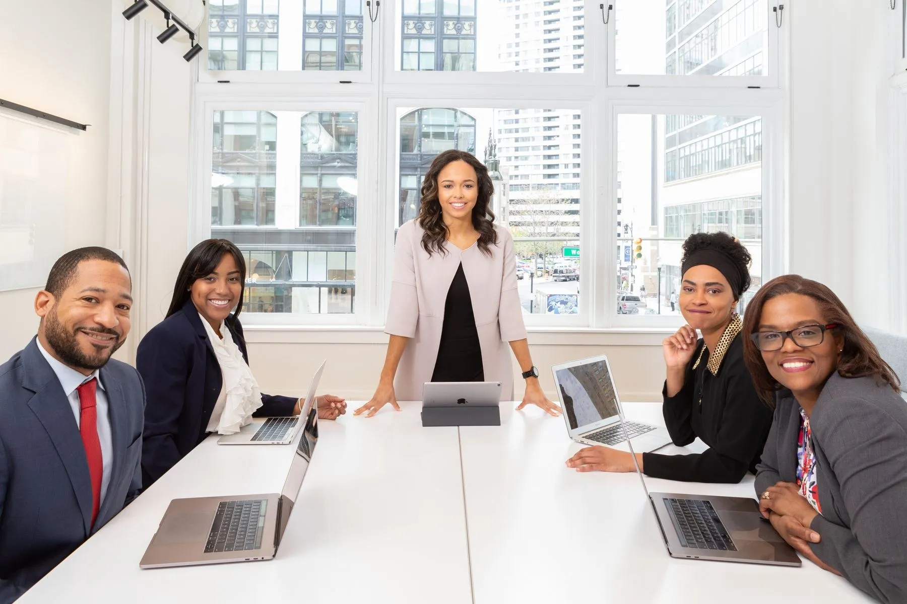 Five colleagues smiling around a bright meeting-room table with laptops, one standing at the head of the table
