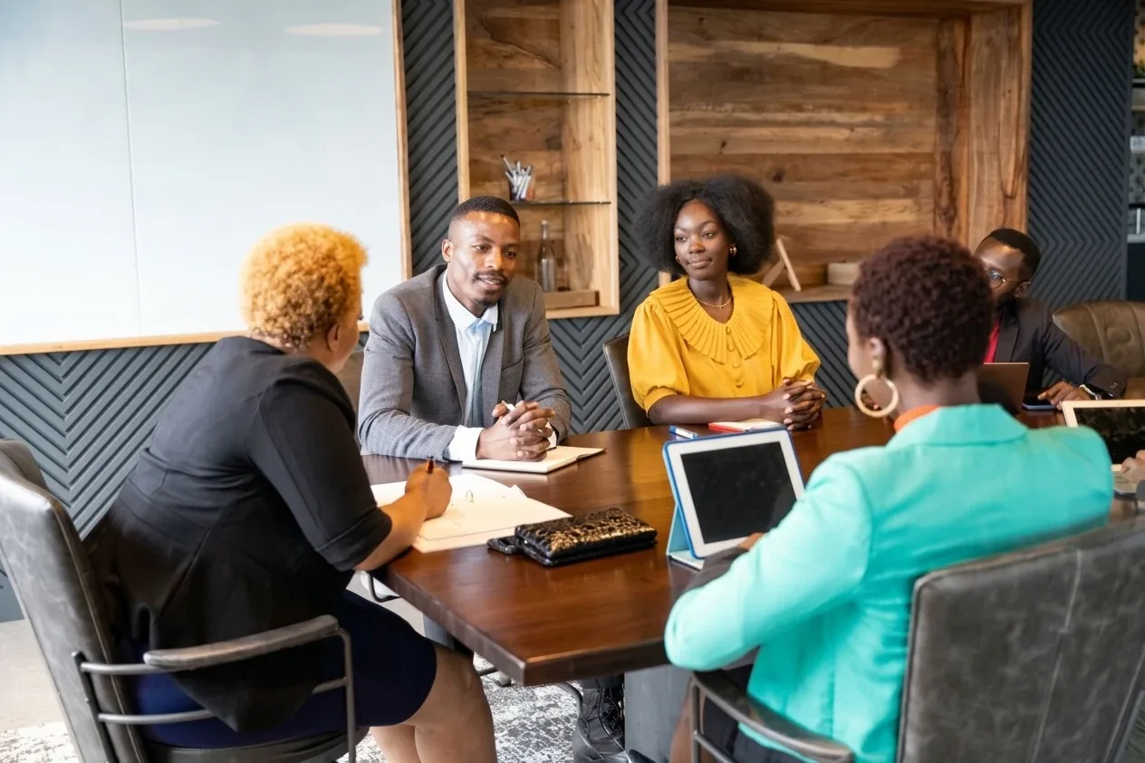 Colleagues meeting around a boardroom table as a man in a grey blazer speaks and a woman in a yellow blouse listens