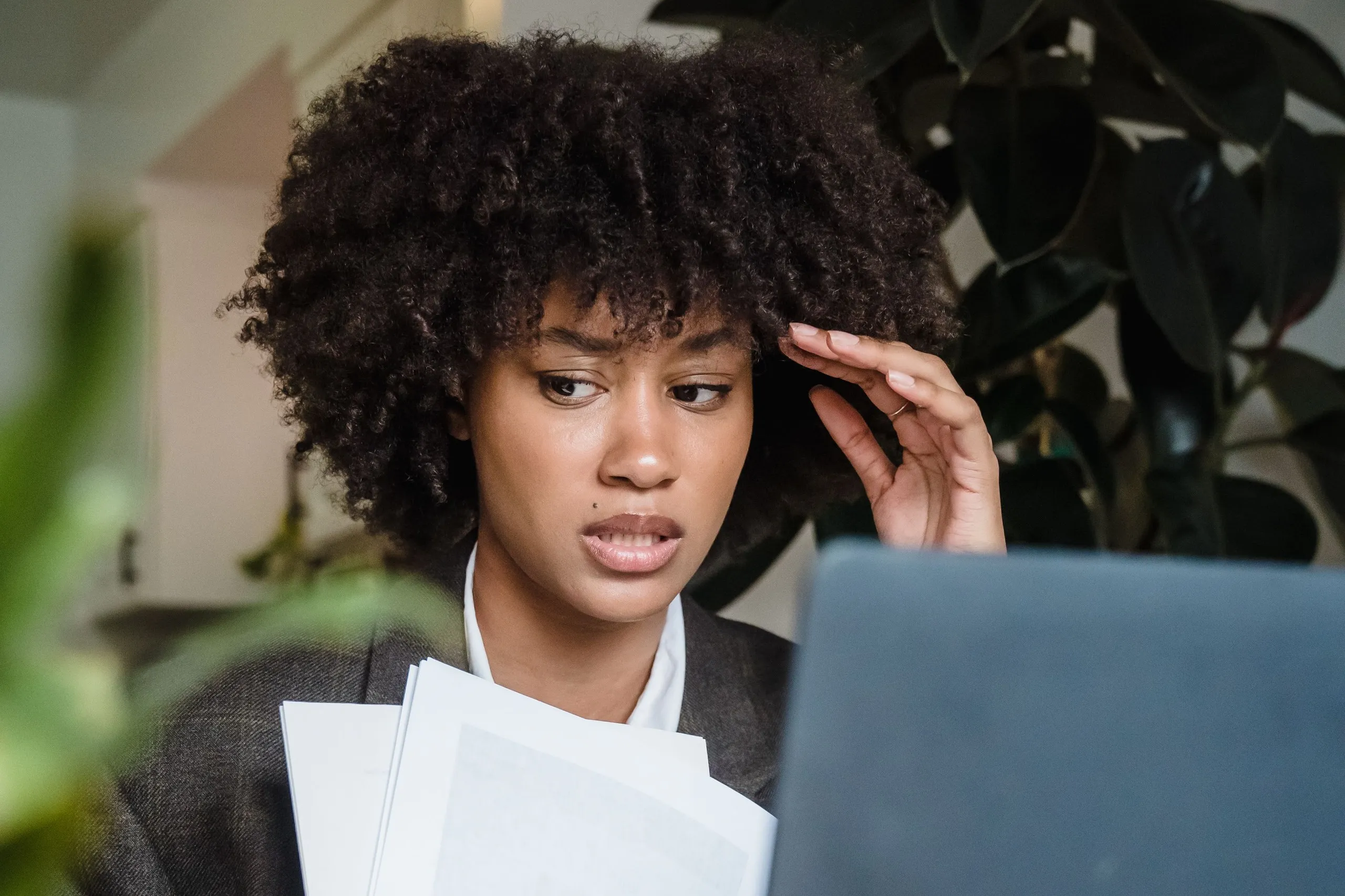 A woman with natural curly hair, in a gray blazer over a white shirt, frowns at an open laptop with her fingertips pressed to her temple, holding a sheaf of printed papers, with a large rubber plant behind her