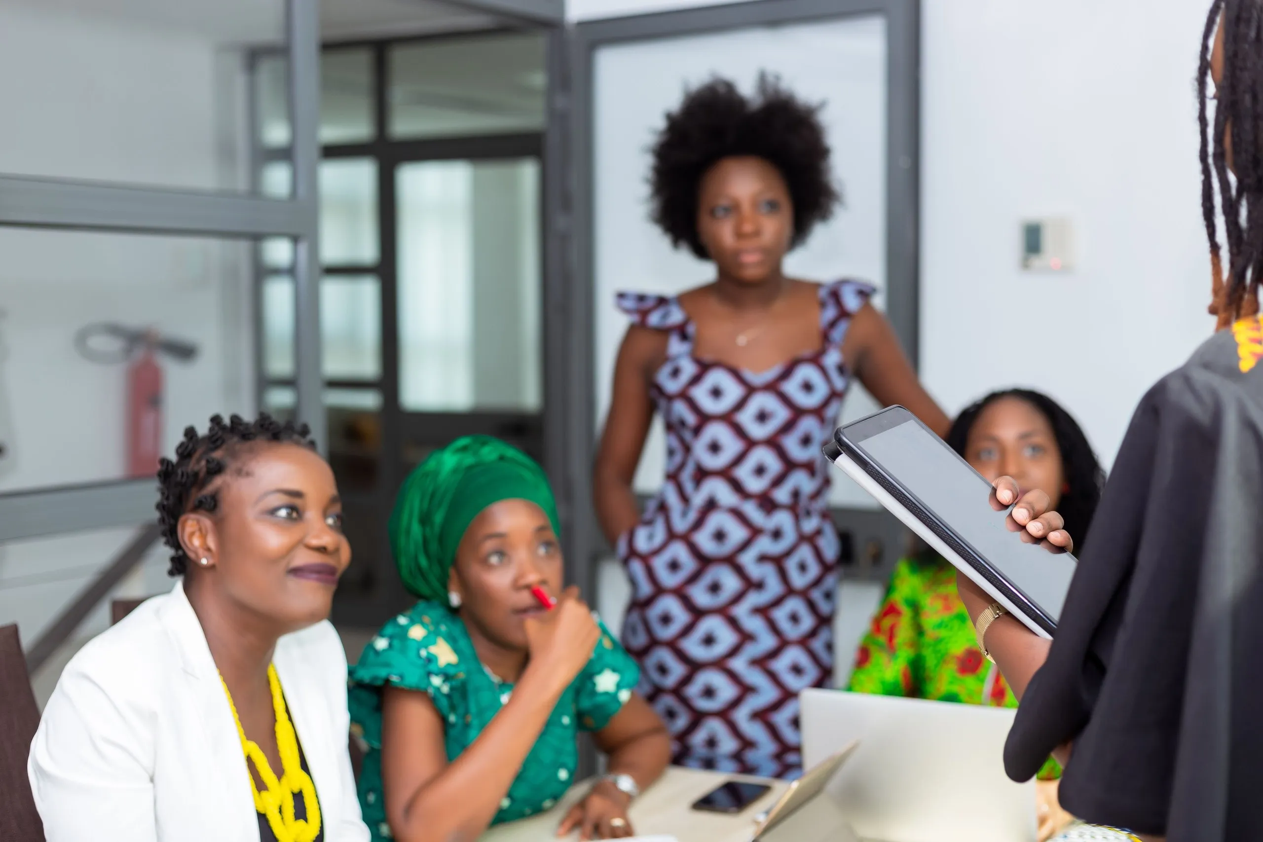 Women at a meeting table listen to a colleague holding a tablet, one in a white blazer and yellow necklace, one in a green head wrap with a pen at her lips, another standing behind them in a patterned dress
