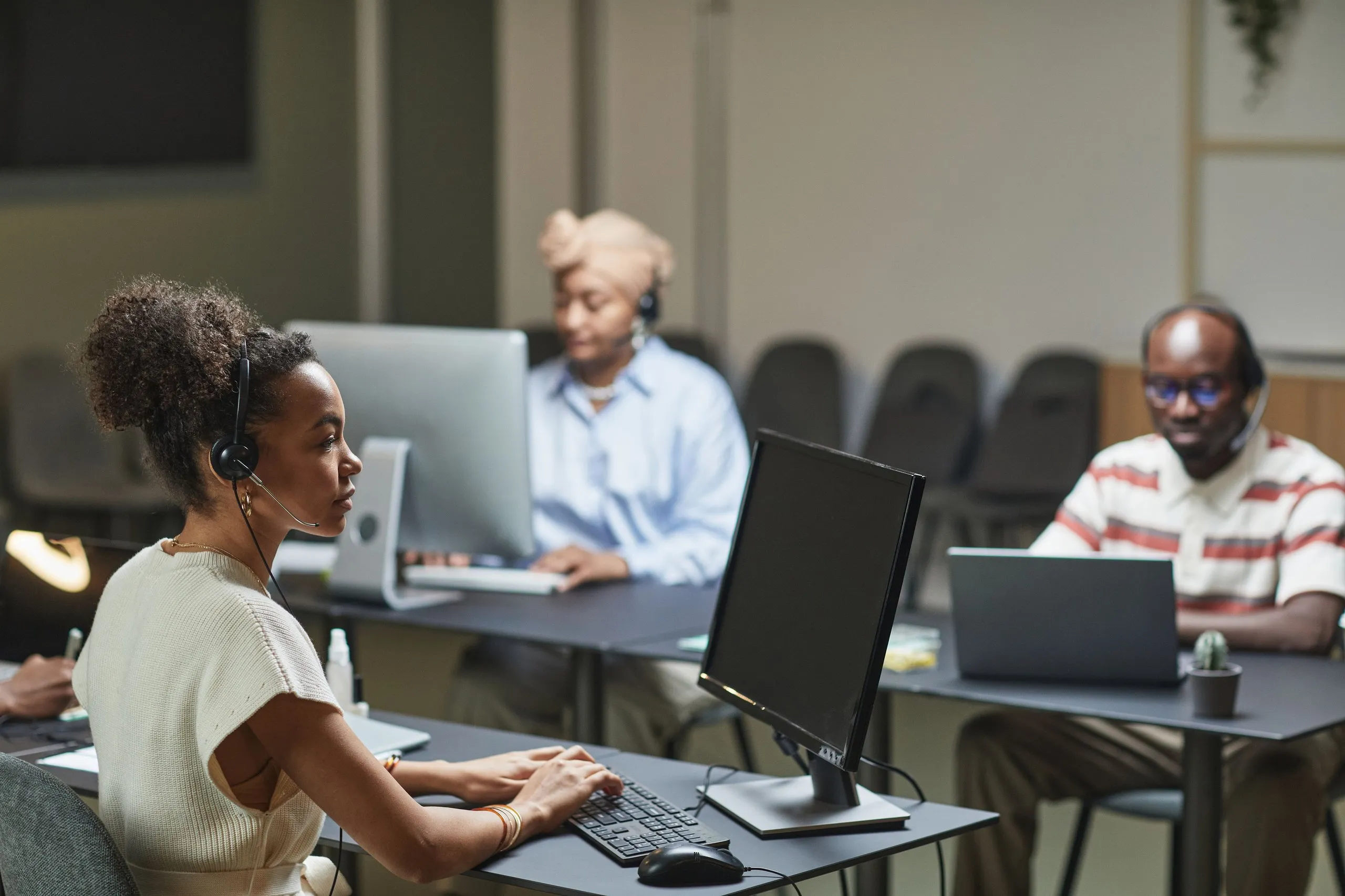 A woman with her curly hair tied up, in a headset and a cream sleeveless knit top, types at a desk beside a monitor while two colleagues in headsets work at desks behind her, a woman in a beige head wrap and pale blue shirt and a man in glasses and a striped shirt