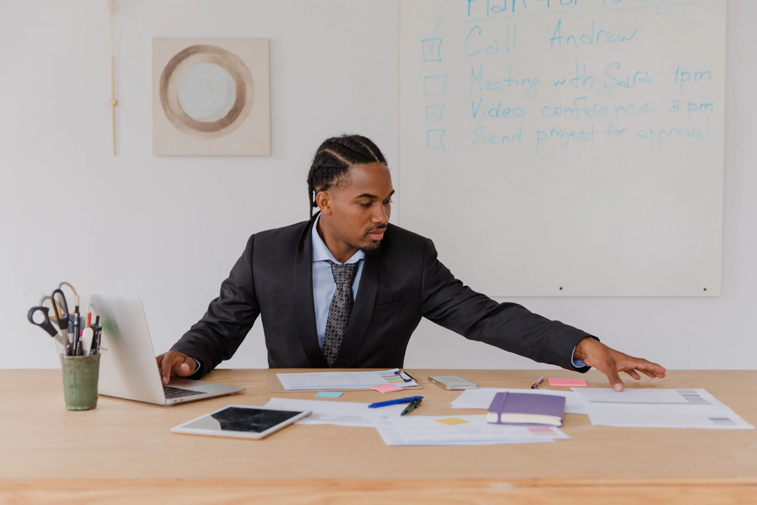 A man in a dark suit and patterned tie sits at a wooden desk covered with papers, sticky notes, a tablet and an open laptop, reaching across to straighten a sheet, in front of a whiteboard listing Call Andrew, Meeting with Sara 1pm, Video conference 3pm and Send project for approval