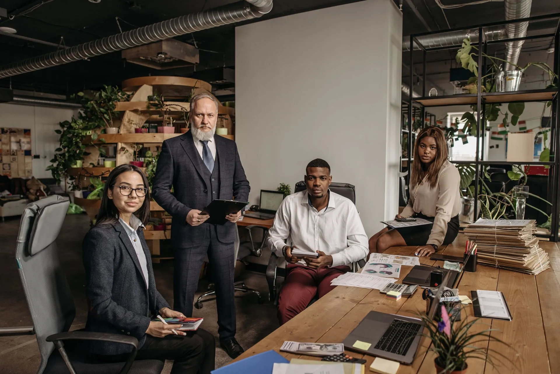 Senior executive with a white beard standing with a clipboard beside three younger colleagues seated at a shared office desk