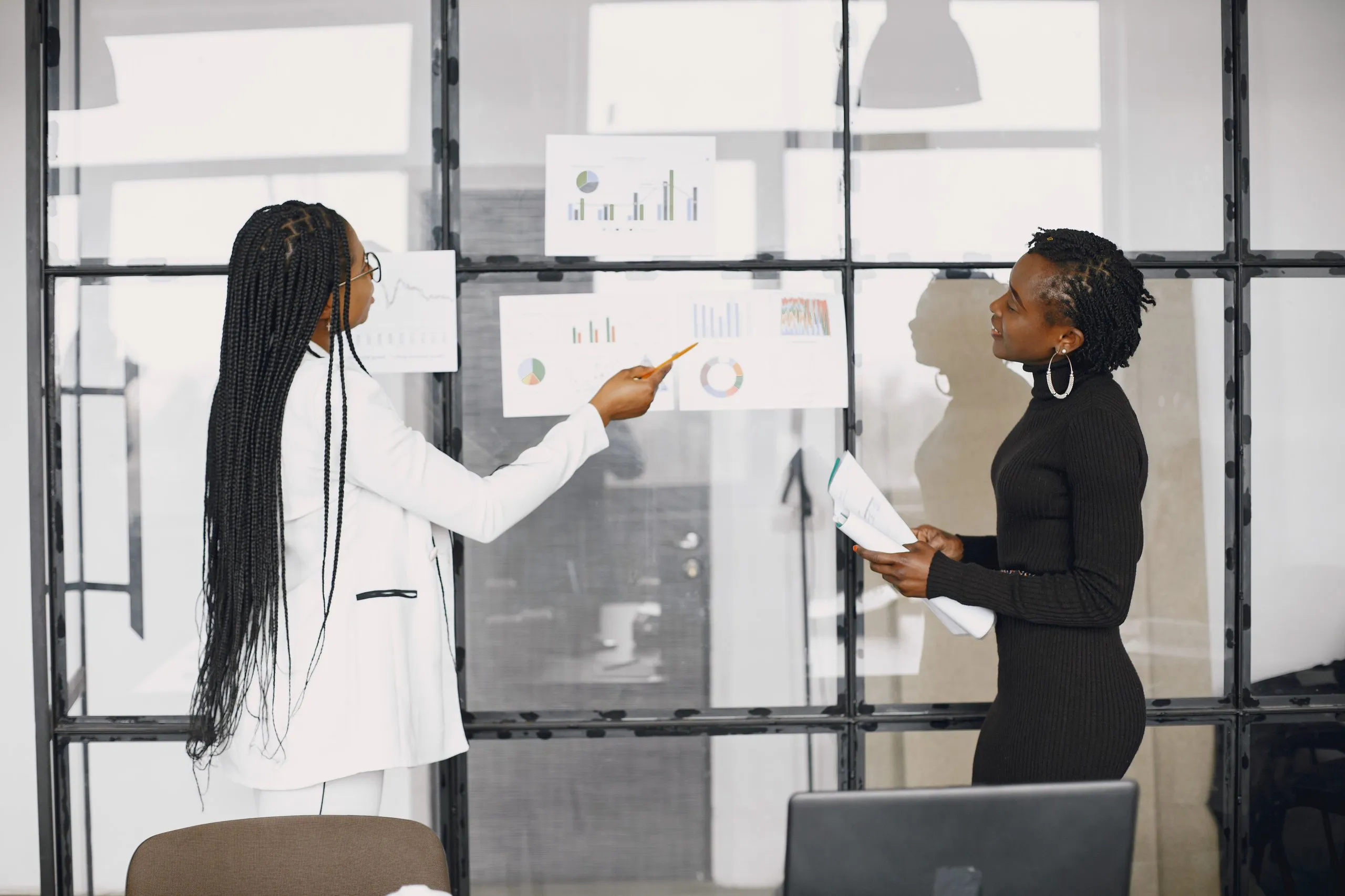 A woman with long braids, glasses and a white blazer points a pencil at bar and donut charts taped to a glass wall, while a colleague in a black turtleneck dress and hoop earrings holds a sheaf of papers and looks on