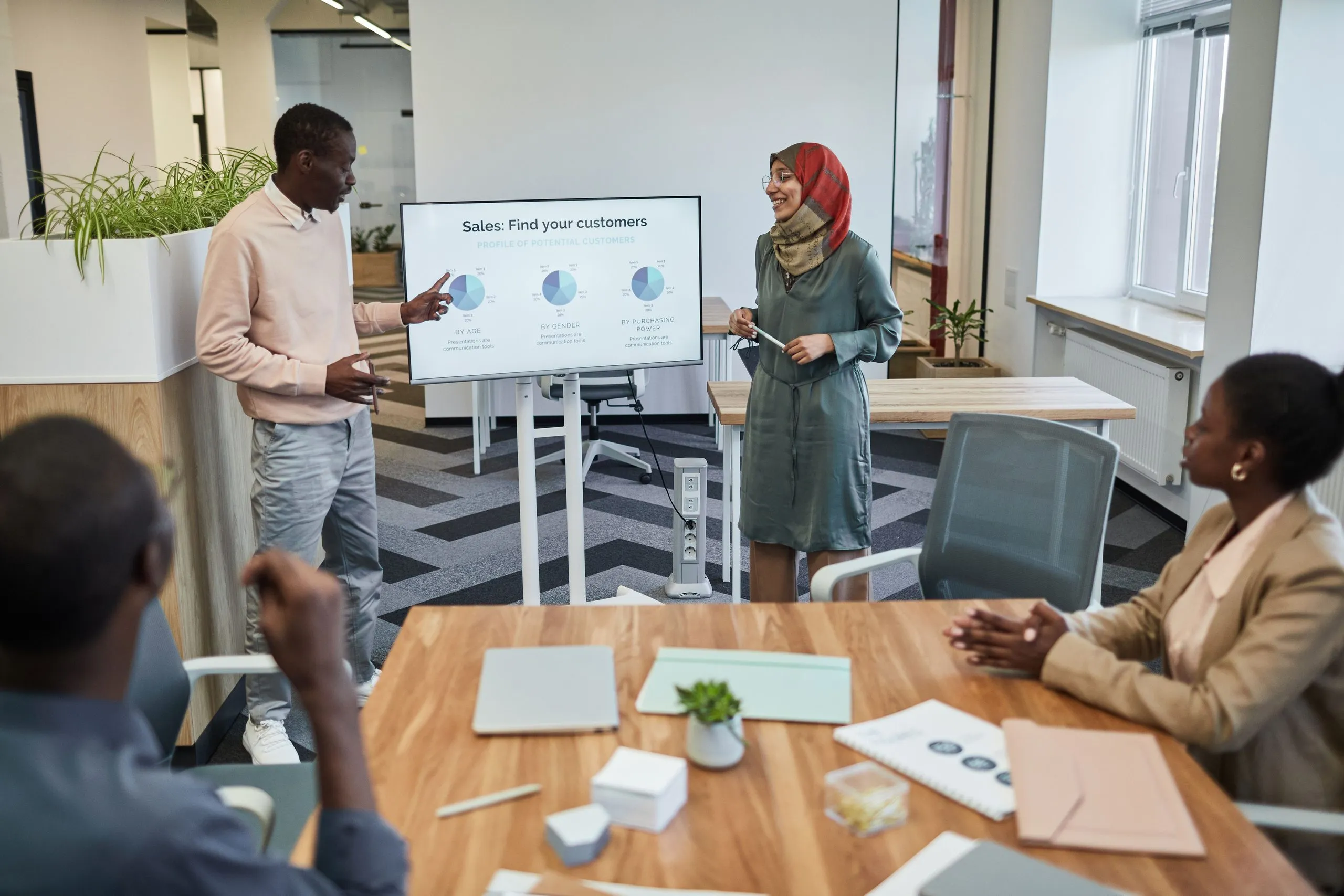 A man in a pink sweater points to a screen of pie charts headed Sales: Find your customers, while a smiling colleague in a red head scarf and green dress stands beside it and two colleagues listen from a wooden meeting table in the foreground