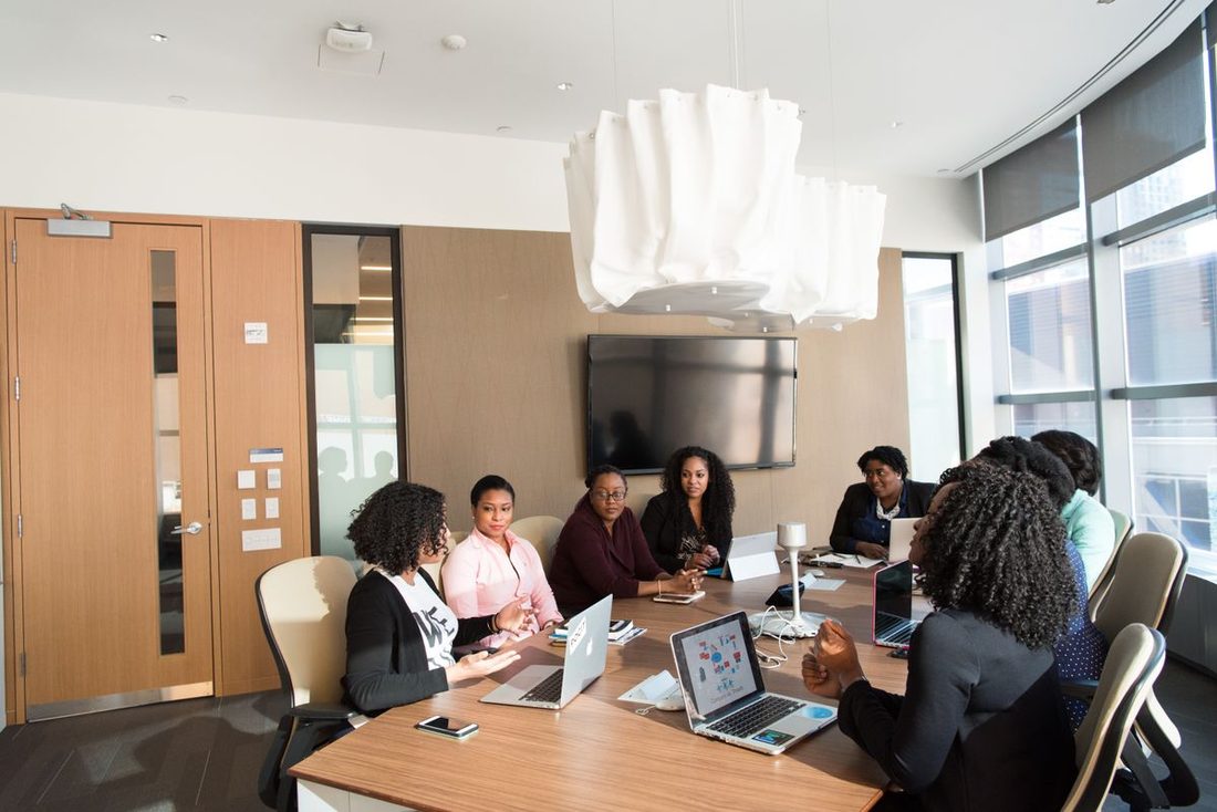 Nine colleagues seated around a long boardroom table with open laptops, one gesturing mid-sentence while the others listen