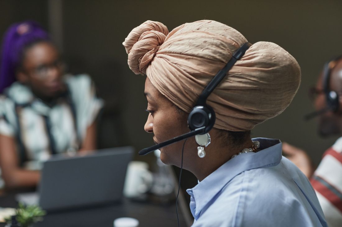 A woman in a peach headwrap, pearl earrings and a pale blue shirt speaks into a headset microphone, seen in profile, while two blurred colleagues in headsets work at laptops behind her