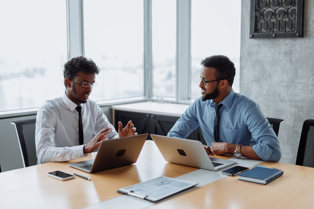 Two colleagues in shirts and ties at a boardroom table with open laptops and a printed report between them, one gesturing as he explains a point to the other