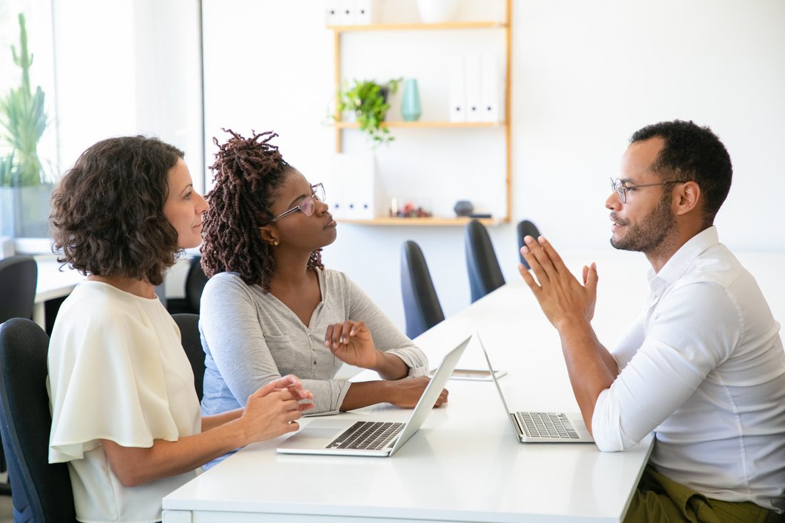 Two women sit side by side at a white table with open laptops, listening closely to a man in glasses and a white shirt across from them who is explaining something with his hands pressed together