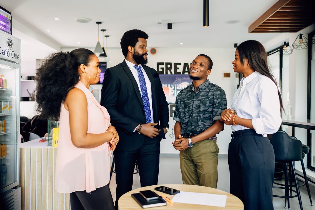 Four colleagues stand talking around a small round table in a bright office cafe: a woman in a pink sleeveless blouse, a bearded man in a dark suit and blue patterned tie, a smiling man in a camouflage-print shirt and a woman in a white shirt, with phones, a notebook and a sheet of paper on the table