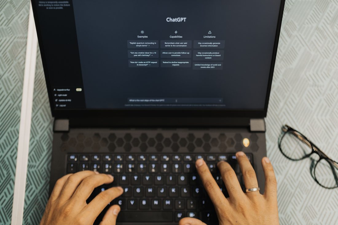 Overhead view of two hands typing on a laptop keyboard, the screen showing ChatGPT's start page with columns headed Examples, Capabilities and Limitations, a pair of glasses resting beside the laptop