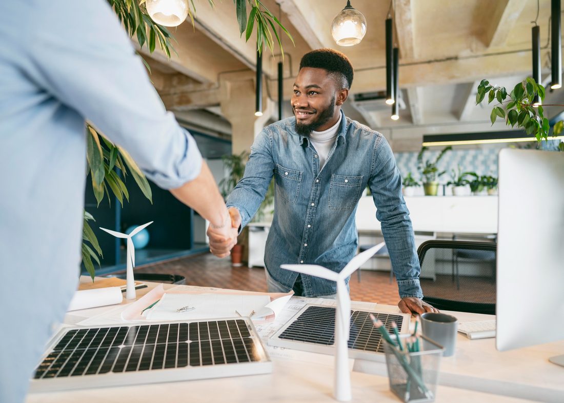 Smiling man in a denim shirt shaking hands across a desk that holds small solar panels, model wind turbines and technical drawings, in an open-plan office with hanging plants