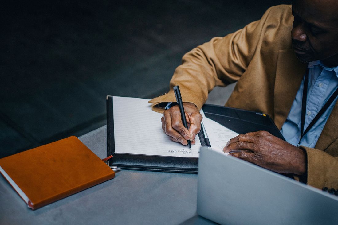 A man in a tan jacket and blue shirt, with a lanyard around his neck, signs his name on a lined page in a black leather folder, beside an orange notebook and an open laptop