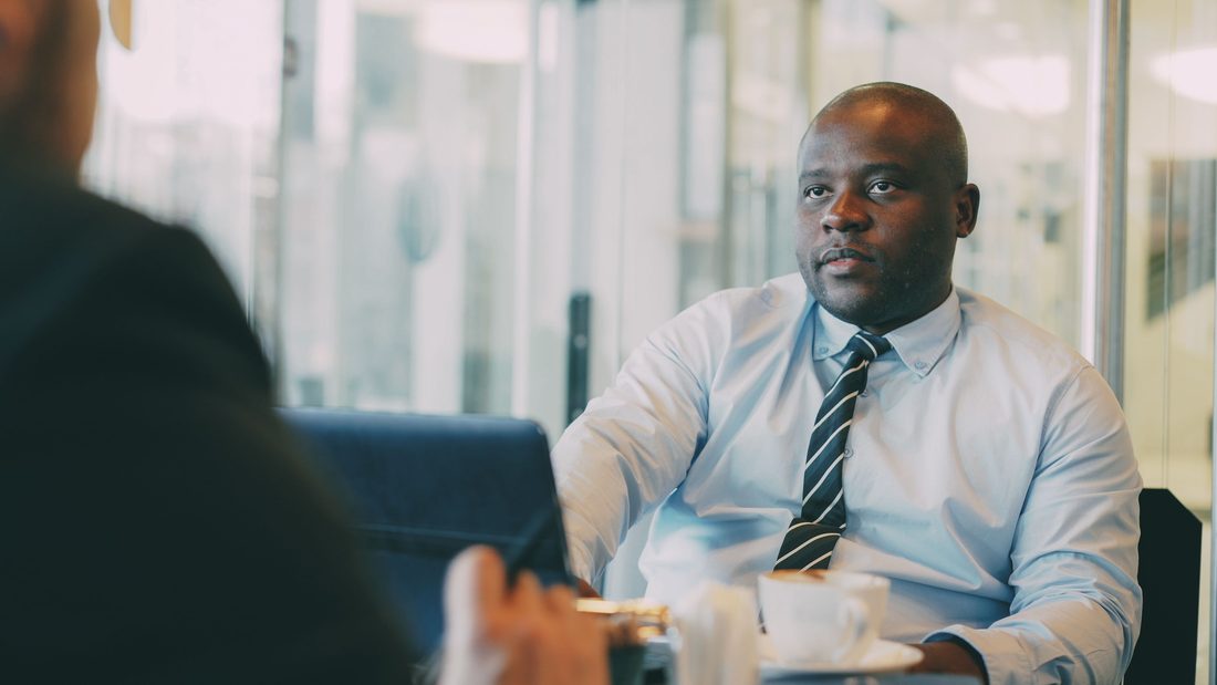 A man in a light blue shirt and striped tie sits at a cafe table with a cup and saucer, listening intently to a colleague whose shoulder and hand are blurred in the foreground, glass walls and soft daylight behind him