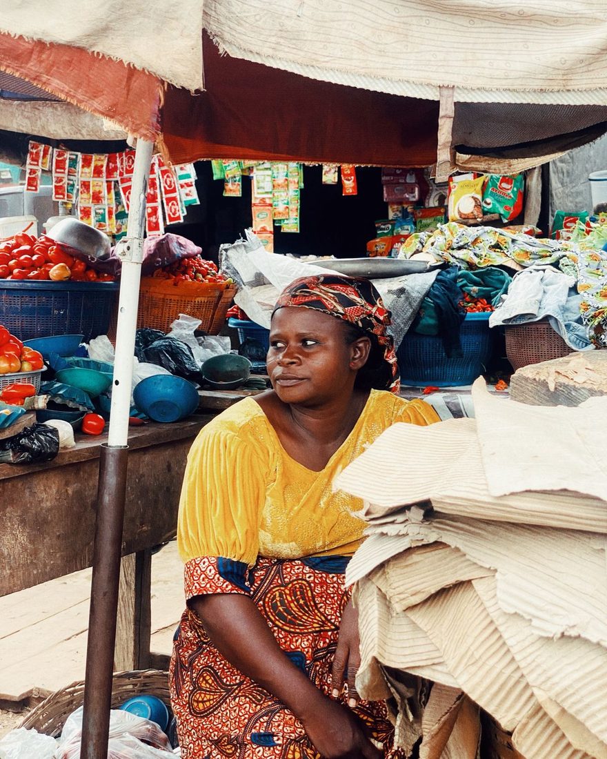 Market trader in a yellow blouse and patterned headscarf seated at her stall between crates of tomatoes, stacked plastic bowls and strings of hanging seasoning sachets