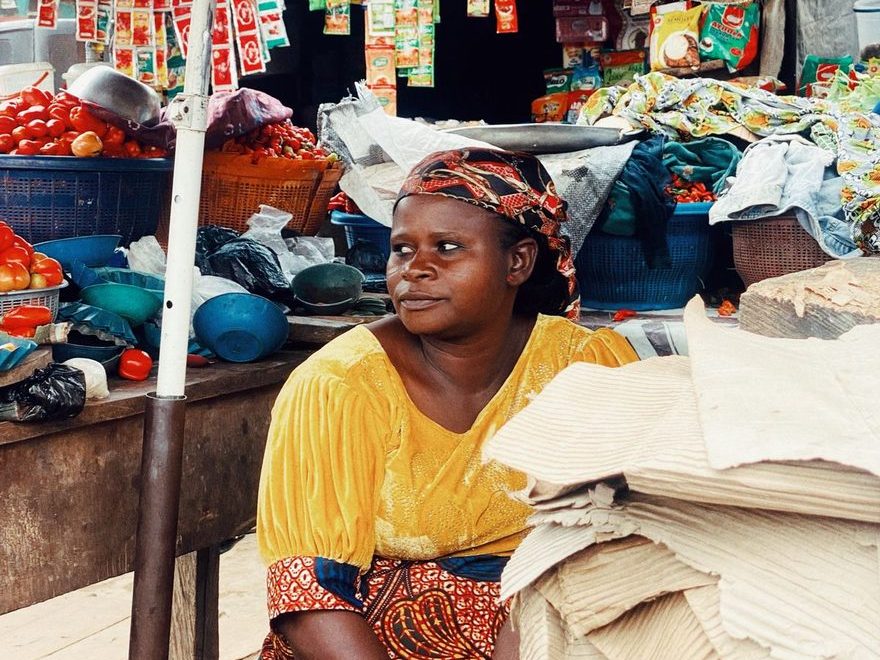 Market trader in a yellow blouse and patterned headscarf seated at her stall between crates of tomatoes, stacked plastic bowls and strings of hanging seasoning sachets