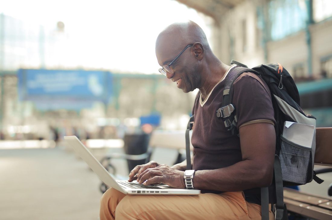 Older man in glasses with a backpack, smiling as he types on a laptop balanced on his knees while seated on a bench in a bright public space