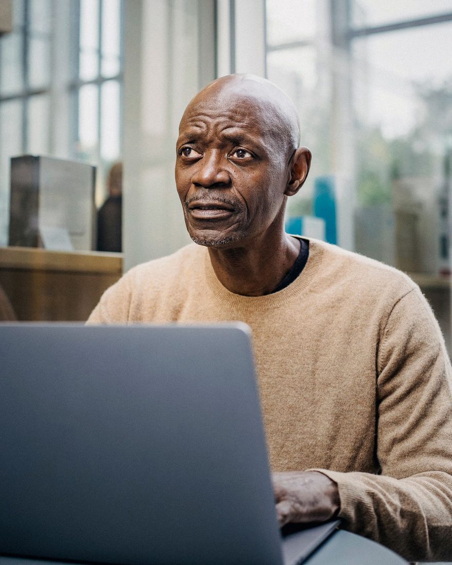Older man in a beige sweater at an open laptop in a glass-walled office, one hand resting on the keyboard, looking away from the screen in thought