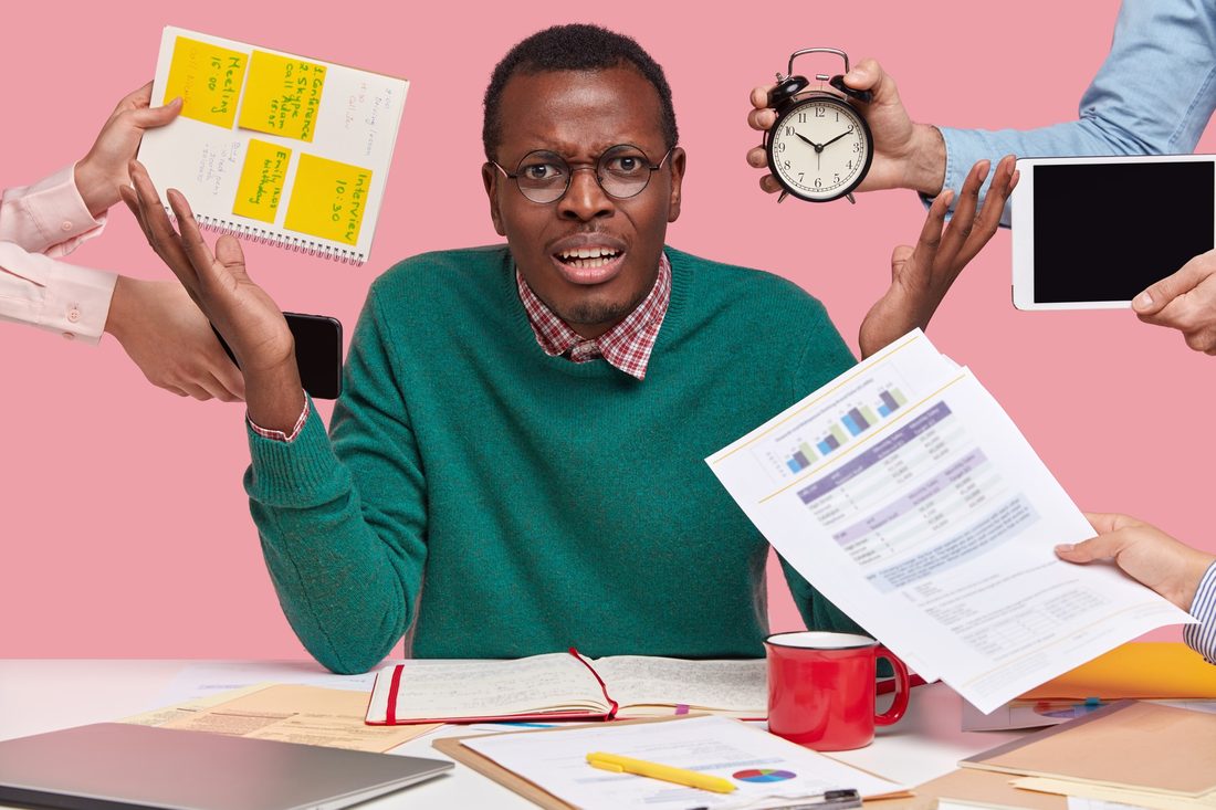 A man in glasses and a green sweater shrugs at his desk, looking overwhelmed, as hands reach in from both sides holding a notebook of sticky notes, a phone, an alarm clock, a tablet and a printed report