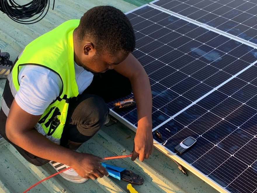 Technician in a high-visibility vest crouching on a corrugated metal roof, crimping an orange cable beside a newly mounted array of solar panels