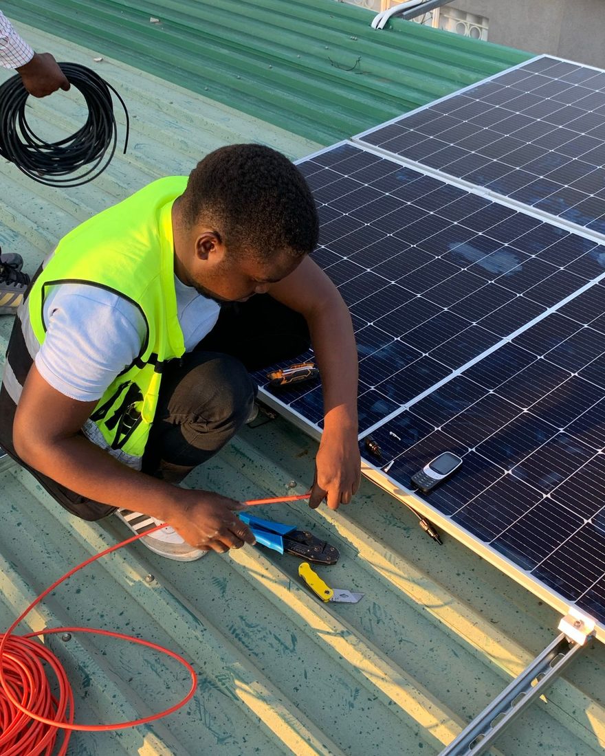 Technician in a high-visibility vest crouching on a corrugated metal roof, crimping an orange cable beside a newly mounted array of solar panels