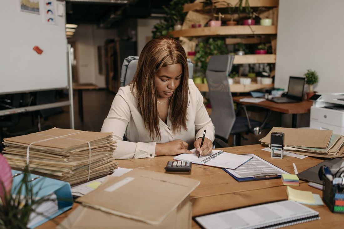 Woman at an office desk writing in a notebook beside a calculator, a clipboard and an ink stamp, with tall stacks of tied brown paper files at her elbow
